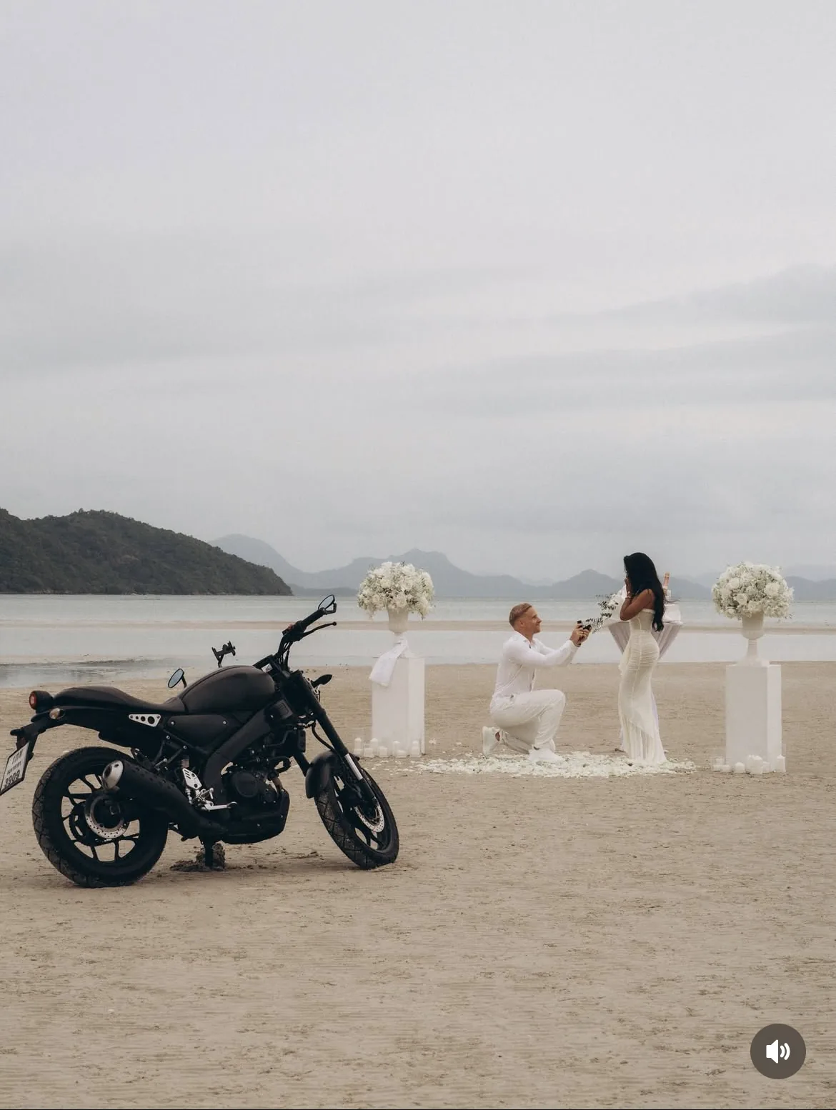 A romantic beach proposal scene in Thailand with a couple beside a motorcycle and white floral arrangements